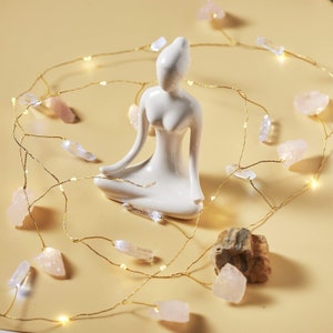 May include: White ceramic figurine of a person sitting in a yoga pose, surrounded by a string of fairy lights with small pink and clear crystal accents.