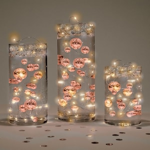 May include: Three clear glass vases filled with water and rose gold coloured glass balls. Each vase has a string of warm white LED lights wrapped around the inside.
