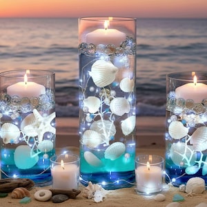 May include: Three clear cylindrical glass vases filled with water, white floating candles, assorted white seashells, faux pearls, and thin strings of glowing LED fairy lights. The water has a blue tint at the base, and the vases sit on sand.