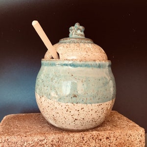 Könnte beinhalten: Ein weißer und blauer Keramik-Honigtopf mit einem Holzrührlöffel. Der Topf hat eine gesprenkelte Glasur und einen dekorativen Deckel.