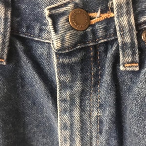 May include: Close-up of the waistband of a pair of blue denim jeans with a metal button that reads "The Cut", with a brown thread seam.