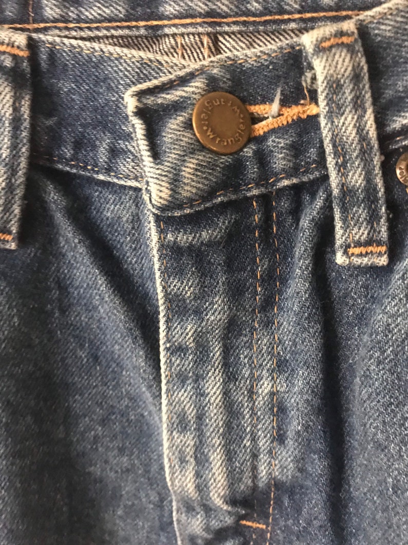 May include: Close-up of the waistband of a pair of blue denim jeans with a metal button that reads "The Cut", with a brown thread seam.