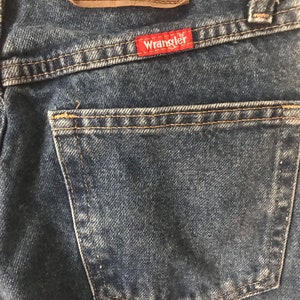 May include: Close-up of a pair of blue denim jeans with a back pocket and a brown leather Wrangler label. The label has the text "Wrangler Authentic Jeans" and "Comfort Superior" printed on it.