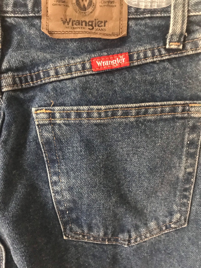 May include: Close-up of a pair of blue denim jeans with a back pocket and a brown leather Wrangler label. The label has the text "Wrangler Authentic Jeans" and "Comfort Superior" printed on it.