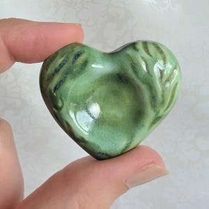 May include: A heart-shaped ceramic object with a glossy, variegated light green glaze. The surface features darker green and blackish accents along the edges and a smooth, concave center, showcasing a reflective finish and organic, flowing color patterns.