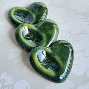 May include: Three heart-shaped ceramic dishes arranged in a diagonal line. Each dish features a glossy, reactive glaze with deep forest green tones transitioning into lighter lime green and dark blue accents, creating a smooth, reflective surface.