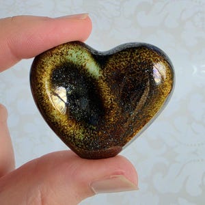 May include: A heart-shaped object with a smooth, glossy finish. The surface features a dark brown and black speckled pattern with a lighter, pale green gradient near the center, creating a reflective, metallic appearance with subtle shimmer.