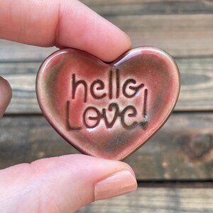 May include: A heart-shaped ceramic ornament with a speckled brown and pink glaze. The ornament has the words "hello love!" written in white.