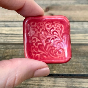 May include: A small, square, pink ceramic dish with a raised, white floral design.