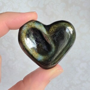 May include: A polished heart-shaped stone featuring a smooth, glossy surface with swirling patterns of deep black, dark green, and iridescent gold hues. The stone displays a reflective finish and a distinct, layered color gradient throughout its form.