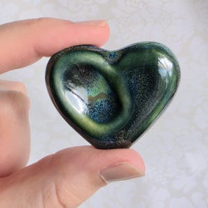 May include: A heart-shaped object with a smooth, glossy finish featuring a blend of deep forest green, dark blue, and black tones. The surface displays a swirling, mottled pattern with reflective highlights across the curved, rounded edges.