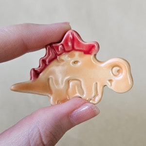 May include: A small ceramic dinosaur figurine with a pink and orange glaze. The dinosaur has a spiky back and a curled tail.