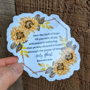 May include: A sticker with a white background and a border of sunflowers. The text on the sticker reads "Now the God of hope fill you with all joy and peace in believing, that ye may abound in hope, through the power of the Holy Ghost. Romans 15:13"