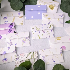 May include: A collection of 10 lavender-themed envelopes with various designs. Some envelopes feature a white background with lavender flowers and green leaves, while others have a purple background with white polka dots or a white background with a lavender floral pattern.