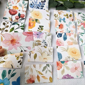 May include: A set of 15 watercolor floral patterned cards. The cards are rectangular and feature various floral designs with pink, yellow, blue, and green colors.