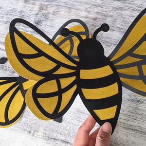 May include: Three black and gold paper bee cutouts. The bee in the center has a black body with gold stripes and wings. The two bees on the sides have gold bodies with black stripes and wings.