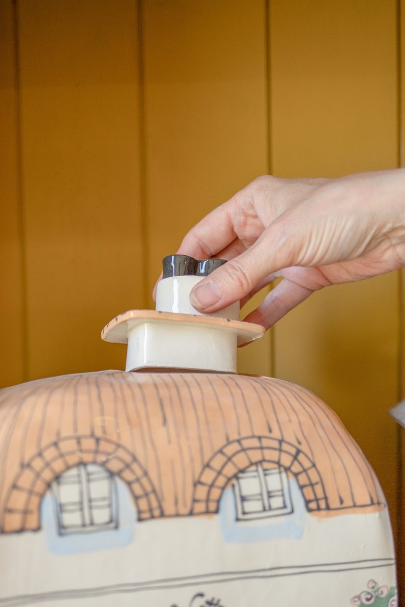 May include: A white ceramic house-shaped cookie jar with a black and white chimney lid. The lid is being lifted by a hand.