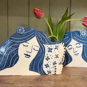 May include: A pair of ceramic vases in the shape of women's faces with long blue hair. One vase holds tulips. The vases are white with blue details. The vase holding the flowers has a floral pattern.
