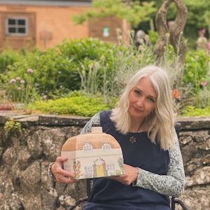 May include: A woman holds a ceramic cottage-shaped bottle with a brown roof and white walls. The bottle has a yellow door and windows and the words "Dove Cottage" are painted on the front.