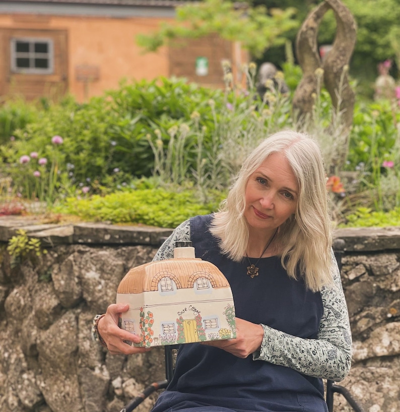 May include: A woman holds a ceramic cottage-shaped bottle with a brown roof and white walls. The bottle has a yellow door and windows and the words "Dove Cottage" are painted on the front.