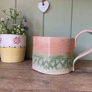 May include: A handcrafted ceramic mug with a curved handle, in pink and green hues. The mug has an embossed pattern around the base. Next to it is a small flower pot with pink flower designs. The mug is suitable for holding 300 ml of liquid.