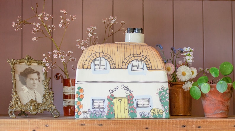 May include: A white ceramic cottage with a thatched roof, painted with a yellow door and windows. The cottage is decorated with flowers and the words "Rose Cottage".