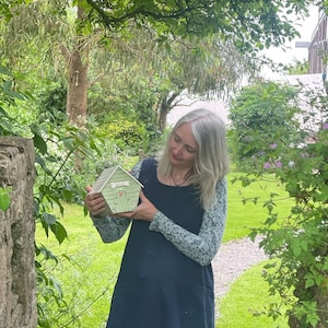 May include: A woman in a blue dress holds a small green model house with a white door and the word "Friend" written on it. The house is surrounded by green foliage.