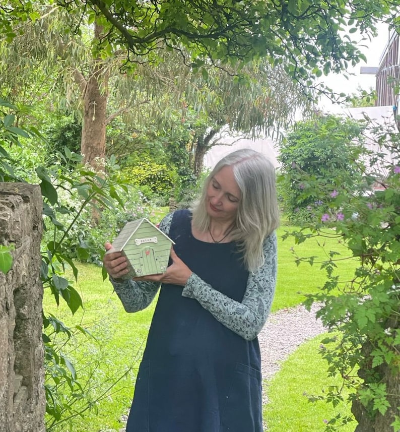 May include: A woman in a blue dress holds a small green model house with a white door and the word "Friend" written on it. The house is surrounded by green foliage.