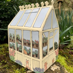 Può includere: Una serra in ceramica dipinta a mano con una cornice giallo chiaro e finestre colorate di blu. L'interno mostra piante in vaso e un gatto. Una volpe e un cane sono dipinti sul davanti. Il tetto ha elementi decorativi.