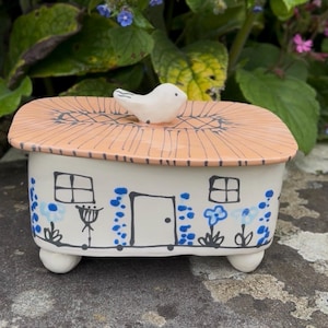 Pode incluir: Uma caixa retangular de cerâmica com uma tampa que apresenta uma pequena figura de pássaro branco no topo. A caixa é branca com contornos pretos pintados à mão de uma casa, incluindo porta, janelas, padrões florais azuis e um telhado cor de pêssego.