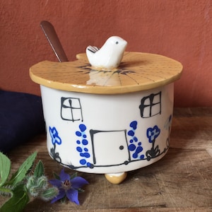 May include: A white ceramic sugar bowl with a bird on the lid. The bowl is decorated with a house design and blue flowers. The bowl has three legs and a spoon rest.