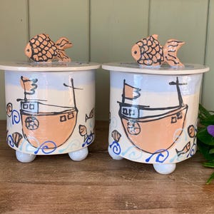 May include: Two ceramic jars with lids, each featuring a hand-painted design of a boat with a fish on top. The jars have a white background with blue and orange accents. The jars are on a wooden surface.