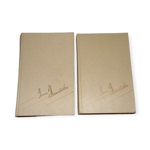 Puede incluir: Dos libros de tapa dura con cubiertas beige se muestran sobre un fondo blanco. Los libros tienen una firma dorada en la esquina inferior izquierda. Los libros están uno al lado del otro, con los lomos hacia la izquierda.