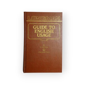 Op de afbeelding: Een bruin hardcover boek met de titel "LONGMAN GUIDE TO ENGLISH USAGE" in gouden letters. Het boek heeft een rechthoekige vorm en staat tegen een witte achtergrond. Het boek bevat ook tekst in het Russisch.