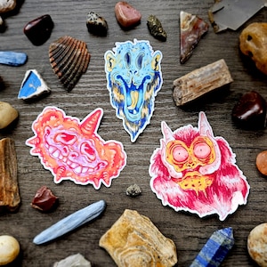 May include: Three colorful monster stickers with unique designs, featuring shades of blue, orange, and red. The stickers are surrounded by various rocks and minerals on a wooden surface.