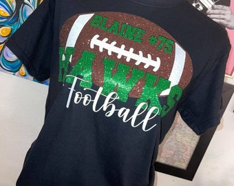 May include: Black t-shirt with a brown and white football design. The football has green glitter and the text "BLAINE #75" and "Football" in white glitter.