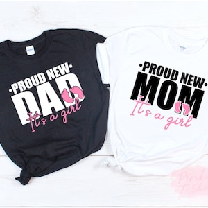 以下が含まれることがあります： 黒と白の2枚のTシャツ。それぞれ「PROUD NEW DAD It's a girl」と「PROUD NEW MOM It's a girl」の文字が白と黒でプリントされています。ピンク色の赤ちゃんの足跡もデザインに含まれています。ベビーシャワー用のTシャツです。