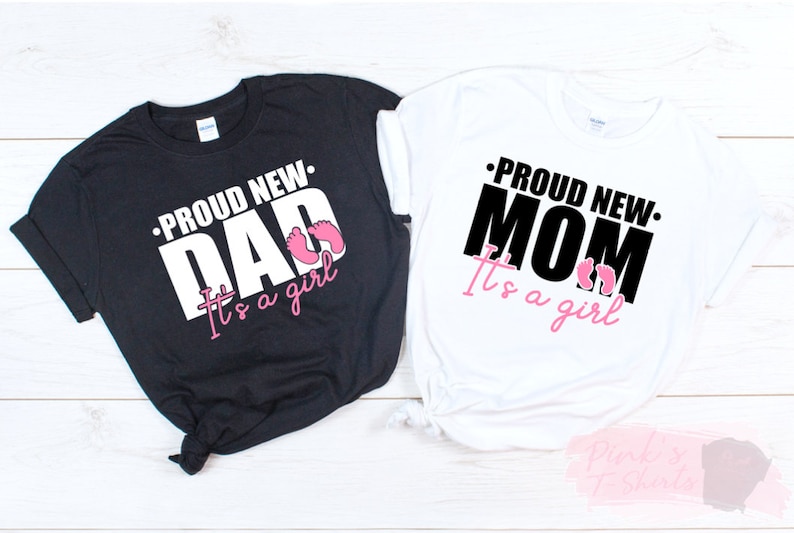 以下が含まれることがあります： 黒と白の2枚のTシャツ。それぞれ「PROUD NEW DAD It's a girl」と「PROUD NEW MOM It's a girl」の文字が白と黒でプリントされています。ピンク色の赤ちゃんの足跡もデザインに含まれています。ベビーシャワー用のTシャツです。