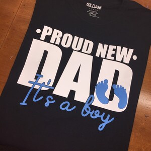 以下が含まれることがあります： 黒いTシャツ。白で「PROUD NEW DAD」、水色で「It's a boy」と書かれています。デザインには青い赤ちゃんの足跡が含まれています。このシャツは柔らかい素材で作られています。