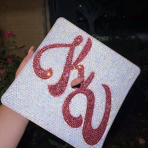 Könnte beinhalten: Eine weiße Abschluss-Kappe, die mit schimmernden Strasssteinen verziert ist und die Buchstaben "KV" in rosa Strasssteinen.