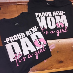 以下が含まれることがあります： 「PROUD NEW DAD It's a girl」と「PROUD NEW MOM It's a girl」と書かれた白い文字がプリントされた2枚の黒いTシャツ。ピンク色の赤ちゃんの足跡もシャツに描かれています。シャツは木製の表面に置かれています。