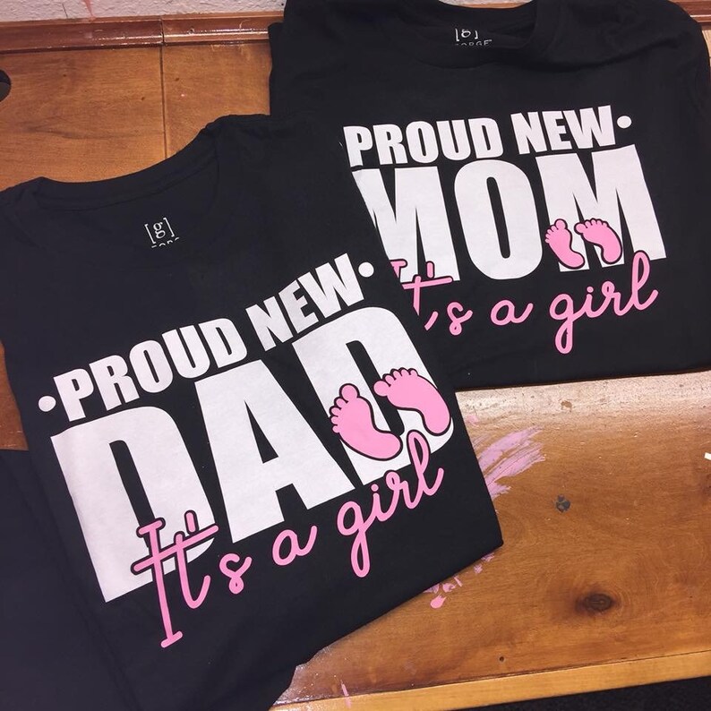 以下が含まれることがあります： 「PROUD NEW DAD It's a girl」と「PROUD NEW MOM It's a girl」と書かれた白い文字がプリントされた2枚の黒いTシャツ。ピンク色の赤ちゃんの足跡もシャツに描かれています。シャツは木製の表面に置かれています。
