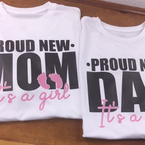 以下が含まれることがあります： 「PROUD NEW MOM」と「PROUD NEW DAD」と書かれた黒い文字がプリントされた2枚の白いTシャツ。テキストの下にはピンク色の赤ちゃんの足跡と、ピンク色の筆記体で「It's a girl」という文字があります。シャツは茶色の表面にあります。