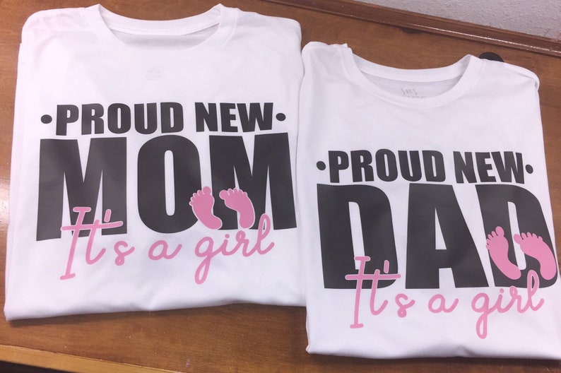 以下が含まれることがあります： 「PROUD NEW MOM」と「PROUD NEW DAD」と書かれた黒い文字がプリントされた2枚の白いTシャツ。テキストの下にはピンク色の赤ちゃんの足跡と、ピンク色の筆記体で「It's a girl」という文字があります。シャツは茶色の表面にあります。