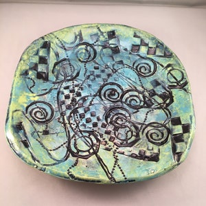 Op de afbeelding: Een keramisch bord met een blauw, groen en zwart geruit patroon en draaiende ontwerpen.