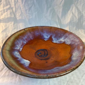 Może przedstawiać: Duża, okrągła ceramiczna miska z brązową glazurą i wzorem białych i fioletowych zwojów. Miska ma niewielki rant wokół krawędzi.