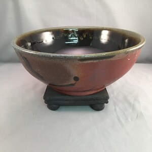 May include: A large, round, ceramic bowl with a black, brown, and red glaze. The bowl is sitting on a small, black wooden stand with three legs.