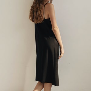 K&ouml;nnte beinhalten: Schwarzes Slip-Kleid mit d&uuml;nnen Tr&auml;gern. Das Kleid reicht bis zur Mitte der Wade und wird mit schwarzen Riemchensandalen kombiniert. Das Model steht vor einer wei&szlig;en Wand.