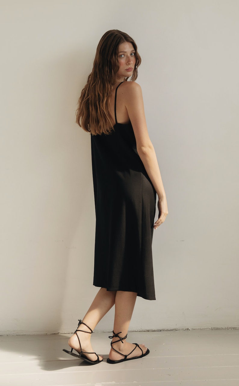 K&ouml;nnte beinhalten: Schwarzes Slip-Kleid mit d&uuml;nnen Tr&auml;gern. Das Kleid reicht bis zur Mitte der Wade und wird mit schwarzen Riemchensandalen kombiniert. Das Model steht vor einer wei&szlig;en Wand.