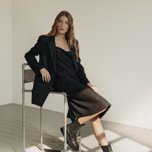 K&ouml;nnte beinhalten: Eine Person tr&auml;gt einen schwarzen Blazer, ein Kleid und Stiefel. Der Blazer hat einen klassischen Schnitt, und das Kleid hat einen einfachen Ausschnitt. Die Stiefel sind schwarz mit einer klobigen Sohle. Die Person sitzt auf einem Metallstuhl in einem Raum mit wei&szlig;en W&auml;nden und Holzboden.
